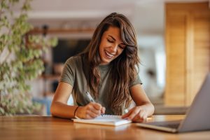 A smiling woman writes a to-do list on a pad of paper. She appears to have no trouble with decision-making.
