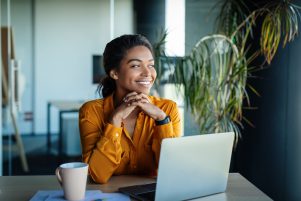 A woman smiles while looking out a nearby window over her left shoulder. Her laptop is open and she has a cup of coffee. She looks relaxed, as if her business is working for her.