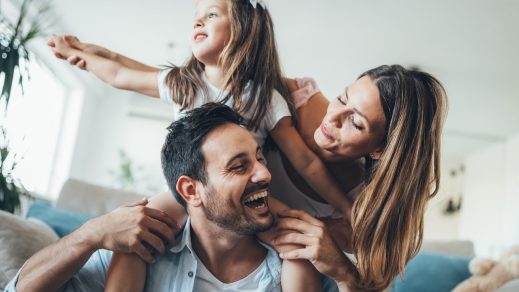 A dad laughs while carrying his young daughter on his shoulders. Mom smiles and looks on. They appear to be full present in the moment.