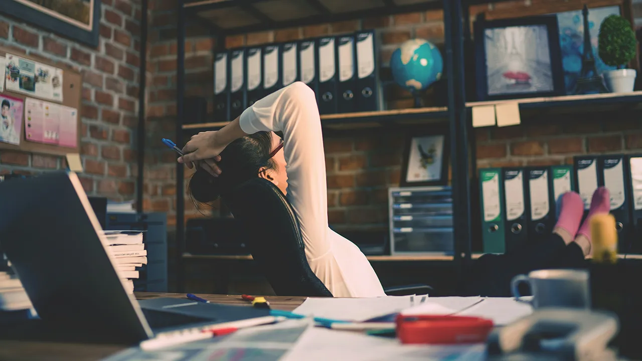 A female entrepreneur slumps back in her chair, her arms clasped over her head as she turns away from her desk, staring into the sky. She appears to be struggling with procrastination and burnout.